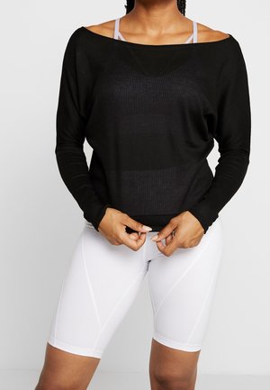 Kobieta ubrana w czarną bluzkę z długim rękawem na lawendowy sportowy biustonosz i białe, sięgające do kolan krótkie spodenki sportowe, ręce poprawiające rant bluzki.