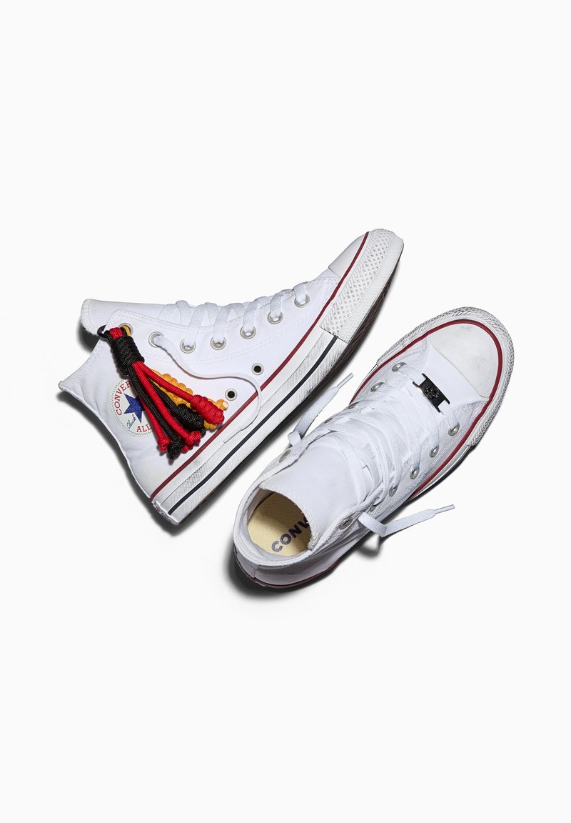 Pair of white high-top Converse sneakers with red and black tassel keychain on left shoe against white background.