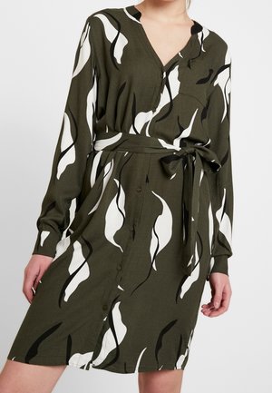 Vrouw draagt een olijfgroene jurk met knopen, lange mouwen, abstracte witte en zwarte bladprint en een strikceintuur in de taille.