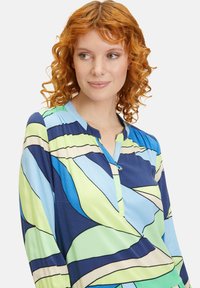 Mehrfarbige Bluse mit blauen, grünen und beigen wellenförmigen Mustern, langen Ärmeln, einem Kragen und Knopfakzenten. Glattes, leichtes Material.