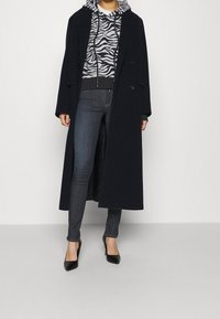 Abrigo largo azul marino con un diseño de doble botonadura sobre una sudadera gris con estampado de cebra, combinado con jeans oscuros y tacones negros con punta.