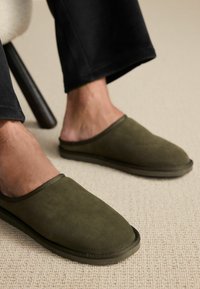 Zöld velúr slip-on klumpák lekerekített orral és texturált felülettel, rugalmas talppal rendelkeznek. Sötét nadrággal viselik egy világos szőnyegen.