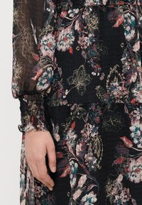 Kvetinové maxi šaty z čierneho materiálu so živými kvetmi, svetlým plisovaním a elastickými manžetami s volánovými okrajmi. Detail rukáva a ruky.