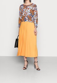 Blusa stampata con maniche lunghe, caratterizzata da un design floreale in blu, arancione e verde; abbinata a una gonna plissettata di lunghezza alla caviglia, di un vivace colore giallo.