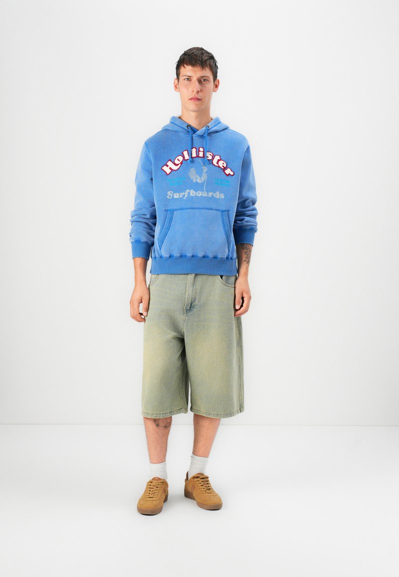 Sudadera con capucha azul con el texto "Hollister Surfboards", combinada con pantalones cortos verdes claros y zapatos marrones, de pie contra un fondo blanco.