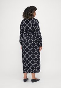 Abito lungo nero con motivo a diamanti bianchi e strisce, maniche lunghe, vita con cintura e orlo alla caviglia. Modella che indossa scarpe nere piatte.