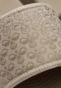 Beige fabric with embossed patterns resembling loops and shapes, featuring a textured surface and contrasting stitched edges.