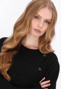 Sort strikket sweater med rund halsudskæring, der har tre brune knapper på venstre skulder og en blød, struktureret overflade.