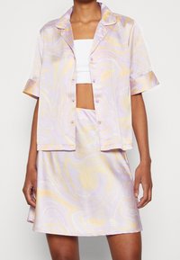 Fialovo-žlutě mramorovaná látka tvoří košili na krátký rukáv s knoflíky a odpovídající sukni, která se vyznačuje hladkou, lesklou texturou a uvolněným střihem.