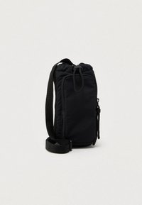 Kompaktowa czarna torebka na ramię z regulowanym paskiem, przednią kieszenią na zamek i zapięciem na sznurek na jasnym, jednolitym tle.