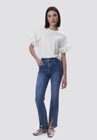 Hvid T-shirt med flæsede stribede ærmer, parret med blå jeans med sidelommer. Model iført sølvfarvede flade sko. Enkel baggrund.