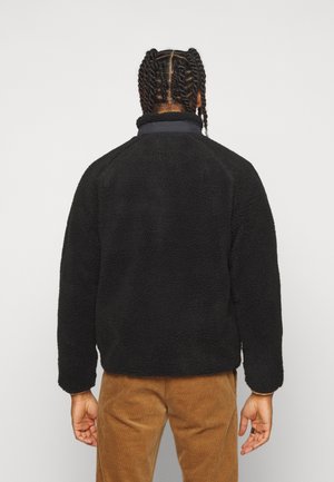 Person mit geflochtenen Haaren, die eine schwarze Fleecejacke und braune Cordhosen trägt, von hinten vor einem schlichten weißen Hintergrund gezeigt.