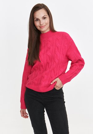 Jasnoróżowy sweter z dzianiny o grubym splotzie, z wysokim kołnierzem i ściągaczami, zestawiony z czarnymi dżinsami. Sweter ma teksturowany, masywny design.