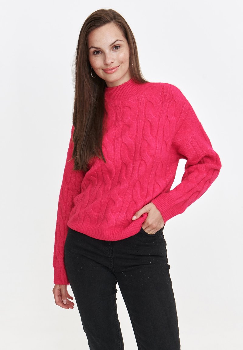 Jasnoróżowy sweter z dzianiny o grubym splotzie, z wysokim kołnierzem i ściągaczami, zestawiony z czarnymi dżinsami. Sweter ma teksturowany, masywny design.