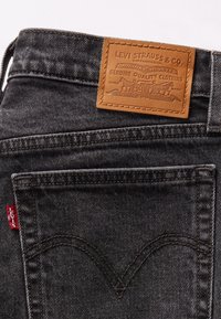 Jean en denim noir avec un patch en cuir présentant un logo et un design embossés. Étiquette de logo rouge près de la poche, surpiqûres contrastantes partout.
