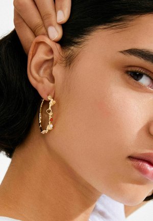 Zlatna naušnica u obliku obruča s teksturiranim dizajnom, ukrašena obojenim perlicama, uključujući narančaste, bijele i prozirne, nošena na uhu modela.