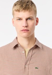 Camisa de cuadros marrón con cuello clásico y logo verde bordado. Tejido suave con un patrón texturizado. Debajo, se ve una parte superior de color beige claro.
