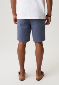 Niebieskie szorty wykonane z lekkiego materiału, z płaskim przodem i luźnym krojem, w połączeniu z brązowymi sandałami na neutralnym tle.