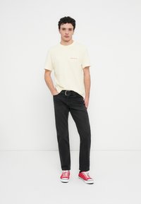 Ανοιχτό κίτρινο βαμβακερό T-shirt με κόκκινο κείμενο, μαύρο τζιν παντελόνι και κόκκινα αθλητικά παπούτσια με λευκές σόλες. Το μοντέλο στέκεται με τα χέρια στις τσέπες.