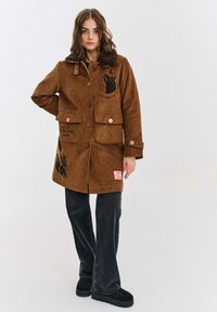 Brązowy płaszcz z korduroju z kołnierzem z sztucznego futra, dwoma przednimi kieszeniami, różowymi guzikami i czarnymi grafikami zwierząt, noszony na ciemnych dżinsach z poszerzanymi nogawkami.