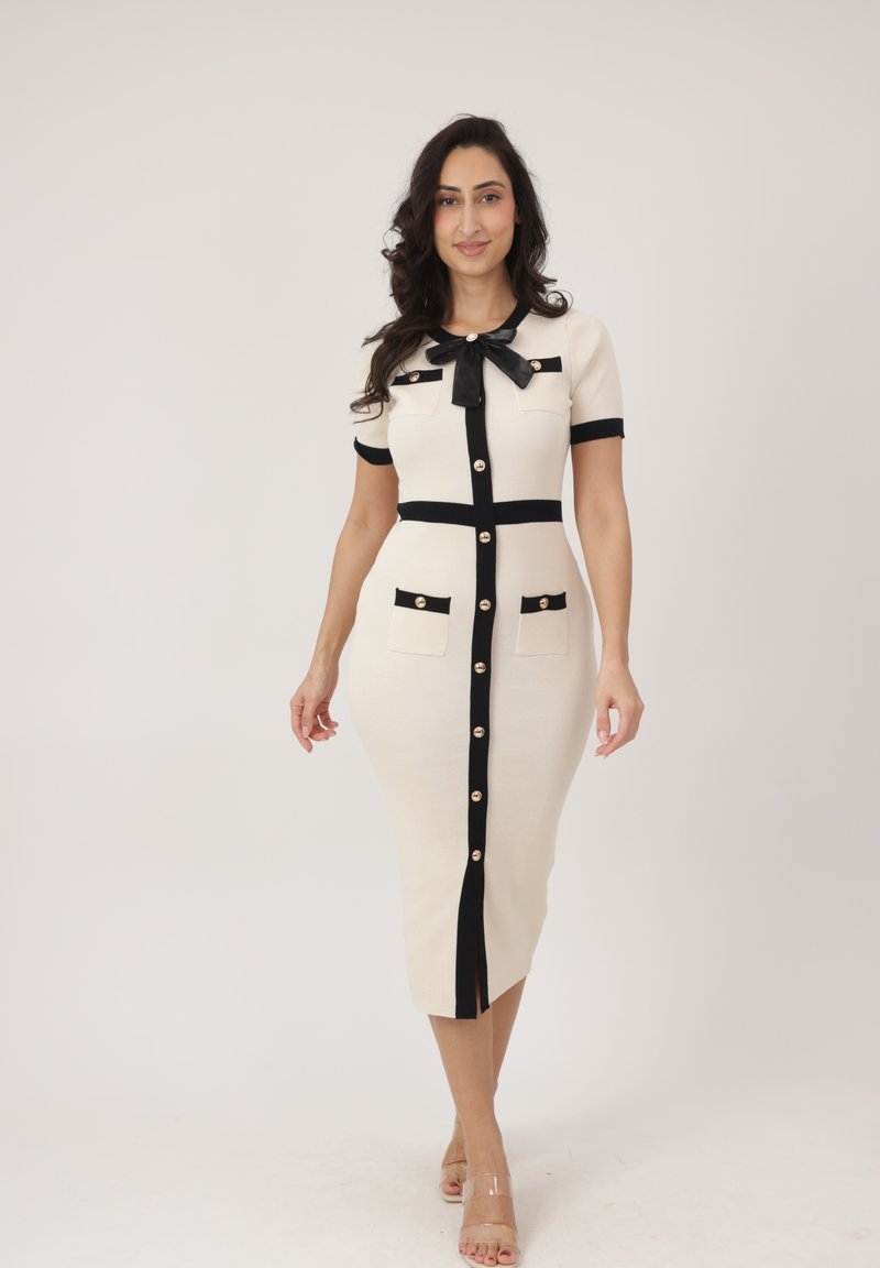 Kobieta ubrana w dopasowaną kremową sukienkę z czarnym wykończeniem, złotymi guzikami z przodu, kokardą przy dekolcie i czterema kieszeniami, idąca na tle jednolitego tła.