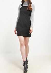 Kvinna iklädd svart hängselklänning över grå långärmad turtleneck, matchat med svarta ankelskor, stående mot vit bakgrund.