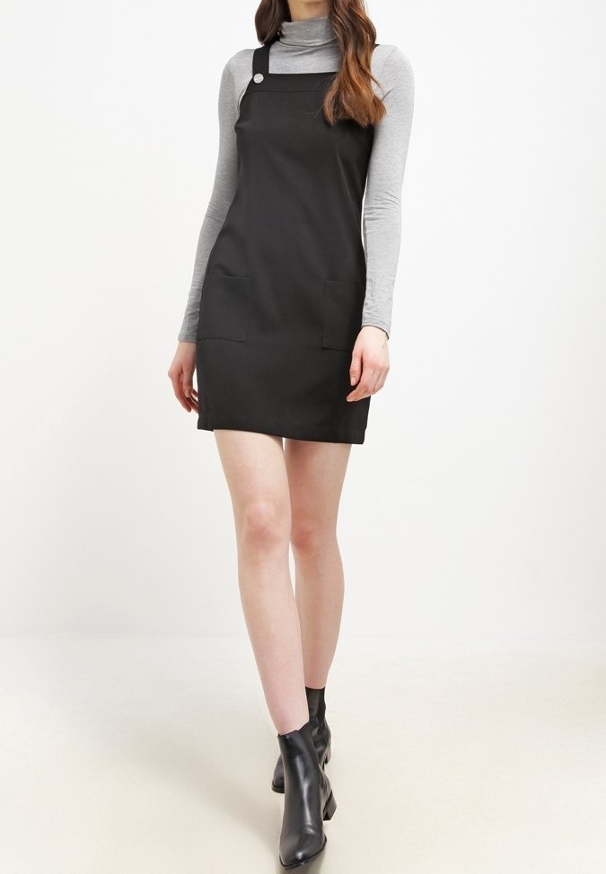 Kvinna iklädd svart hängselklänning över grå långärmad turtleneck, matchat med svarta ankelskor, stående mot vit bakgrund.