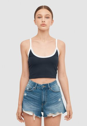 Joven mujer de pie, con postura recta, vistiendo un crop top negro con ribete blanco y pantalones cortos vaqueros azules desgastados, sobre un fondo liso.