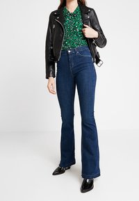 Černá kožená motorkářská bunda přes zelenou květinovou blůzu, v kombinaci s vysokými modrými zvonovými džíny a černými lesklými kotníkovými botami.