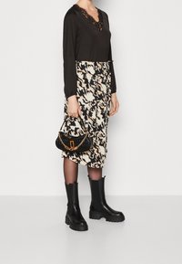 Camiseta negra de manga larga con escote en forma de V de encaje, falda beige y negra con estampado, botas de goma negras hasta la rodilla y un bolso negro texturizado con cadena.
