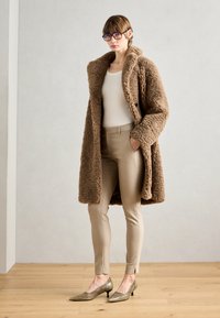 Brun kunstig pelsjakke med sjal-krage, beige tettsittende bukser og metalliske lave hæler. Modellen står på tregulv mot en lys vegg.