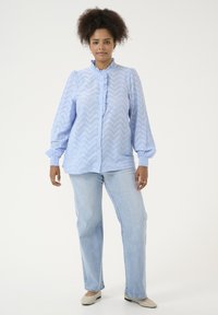 Chemise bleu clair à boutonnières avec un col en volants et des manches longues, arborant un motif en chevrons et des poignets élastiques. Portée avec un jean bleu clair.