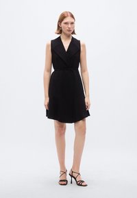 Μαύρο αμάνικο φόρεμα με V-λαιμόκοψη, εφαρμοστή μέση και φούστα που ανοίγει. Λείο ύφασμα με μινιμαλιστικό σχέδιο χωρίς ορατά μοτίβα ή διακοσμητικά στοιχεία.