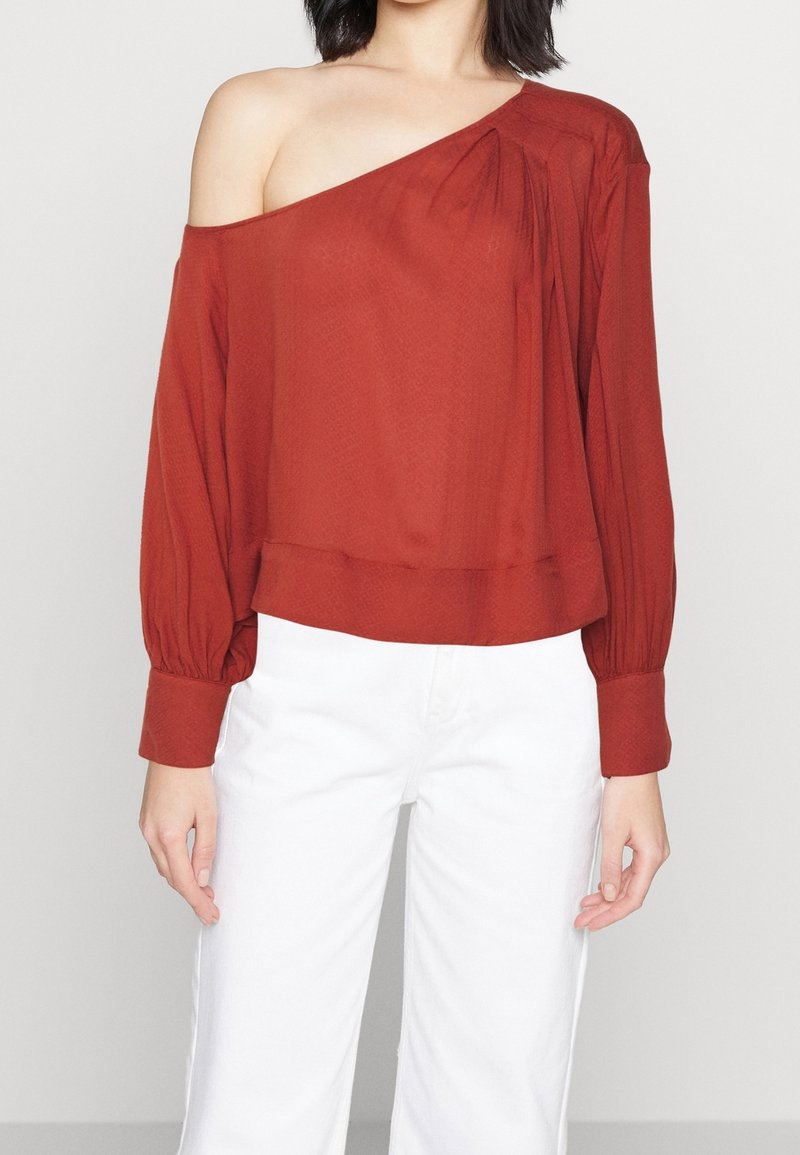 Femme portant un chemisier à manches longues couleur rouille, décolleté épaules, associé à un pantalon blanc taille haute sur un fond uni.
