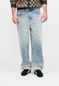 Lichtblauwe spijkerbroek met een vervaagd, overal aanwezig logo-patroon. Rechte pijpen, vijfzakkenmodel, gedragen met zwarte sneakers.