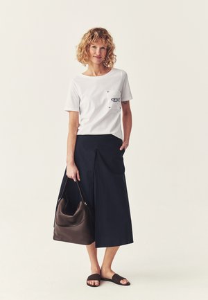 Vrouw met krullend haar, die een wit overhemd met vismotief draagt, een marineblauwe midirok, bruine sandalen, en een grote bruine handtas vasthoudt.