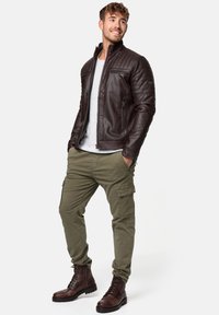 Chaqueta de cuero marrón con hombros acolchados, suéter gris debajo, pantalones cargo caquis y botas marrones con cordones. Diseño moderno y bien ajustado.