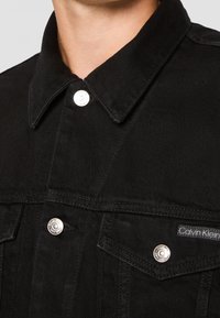 Svart denimjacka med spetsig krage, silverfärgade tryckknappar, två bröstfickor och en Calvin Klein-logotagg på fickan.