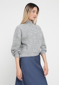 Pulover gri tricotat cu guler înalt, model texturat și mâneci bufante, purtat cu o fustă albastră din satin.