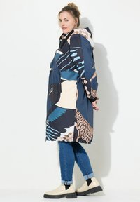 Cappotto da pioggia lungo e colorato con motivi di uccelli e astratti, realizzato in materiale resistente all'acqua, dotato di cappuccio e tasche con zip.