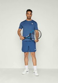 Blå atletisk t-shirt med en logotyp, matchande blå shorts, håller en tenniskö. Vita skor och strumpor fulländar outfiten.