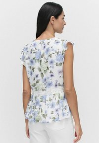 Blumenbluse mit kurzen, gefransten Ärmeln, die blaue und grüne Blumen auf einem weißen Hintergrund zeigt, und einer Spitzenakzent an der Taille.