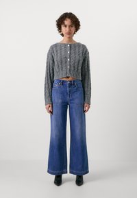 Pull en maille torsadée gris, court, avec encolure ronde et fermeture à boutons, associé à un jean bleu à jambes larges et des bottines noires à bout pointu.