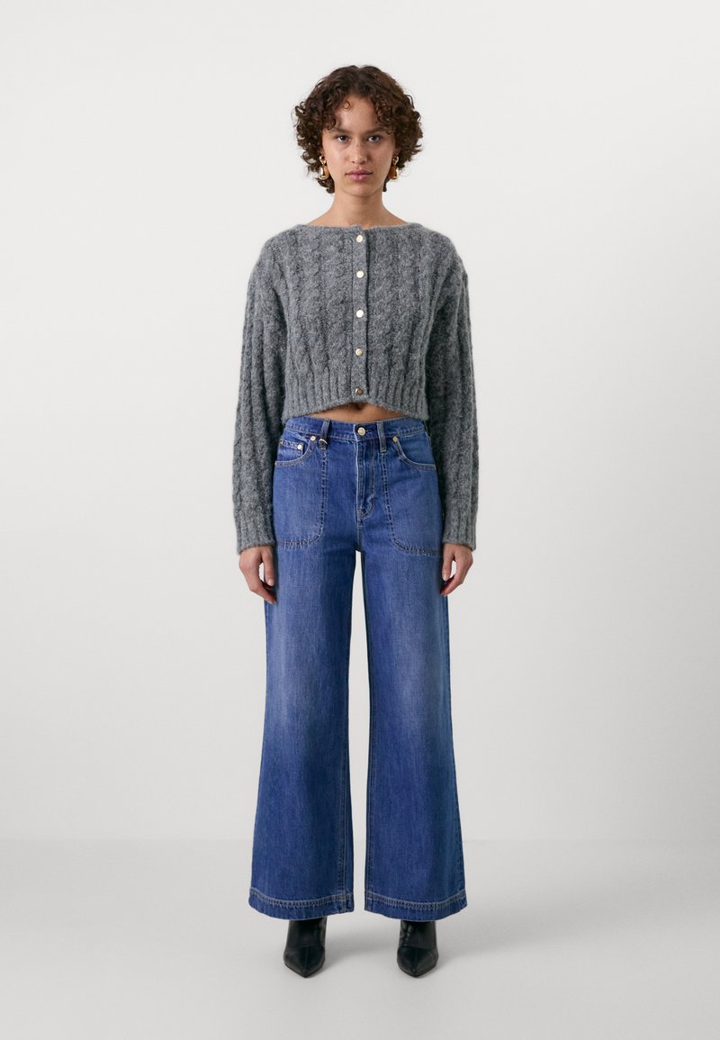 Pull en maille torsadée gris, court, avec encolure ronde et fermeture à boutons, associé à un jean bleu à jambes larges et des bottines noires à bout pointu.