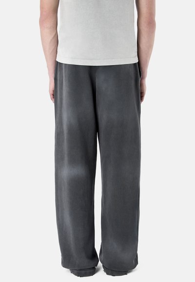 Personne portant un pantalon ample gris foncé et une chemise gris clair, vue de dos sur un fond clair uni.