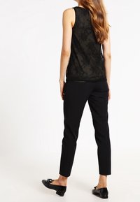 Mujer de pie con una blusa sin mangas de encaje negro, pantalones negros ajustados y mocasines negros con borlas, de espaldas sobre un fondo blanco.