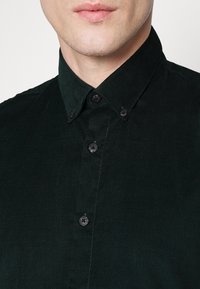 Tummanvihreä corduroy-paita, jossa on klassinen kaulus. Paidan etuosassa on mustia nappeja ja teksturoitu pinta. Lähikuvassa paidan yläosa.