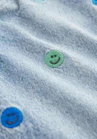 Tessuto in denim blu chiaro con faccine sorridenti ricamate in verde e blu, che aggiungono texture e interesse visivo al materiale.