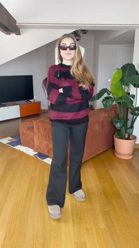 Ριγέ μπορντό και μαύρο πουλόβερ, μαύρο παντελόνι και γκρίζα παντόφλα. Μακριά μαλλιά, γυαλιά ηλίου. Δωμάτιο με φυτό και καφέ καναπέ. Ξύλινο πάτωμα.