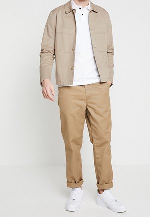 Mężczyzna ubrany w beżową kurtkę, białą koszulkę polo, beżowe spodni z mankietem i białe sneakersy na tle jednolitego białego tła.